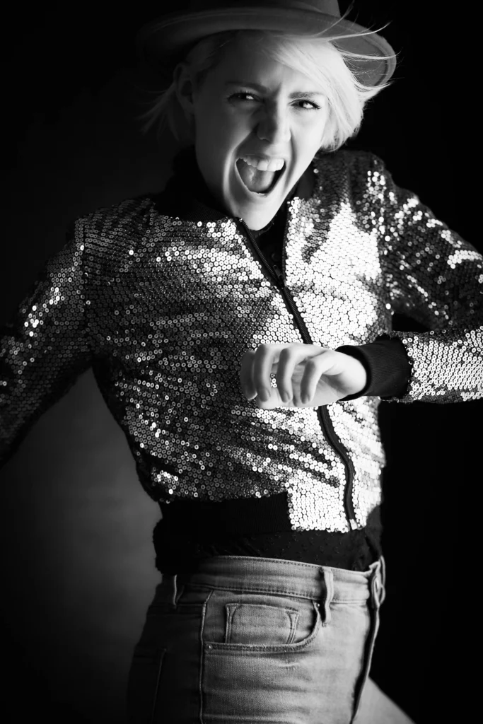 black and white image of a confidently exclaiming woman wearing a sequin jacket, felt brimmed hat and jeans.