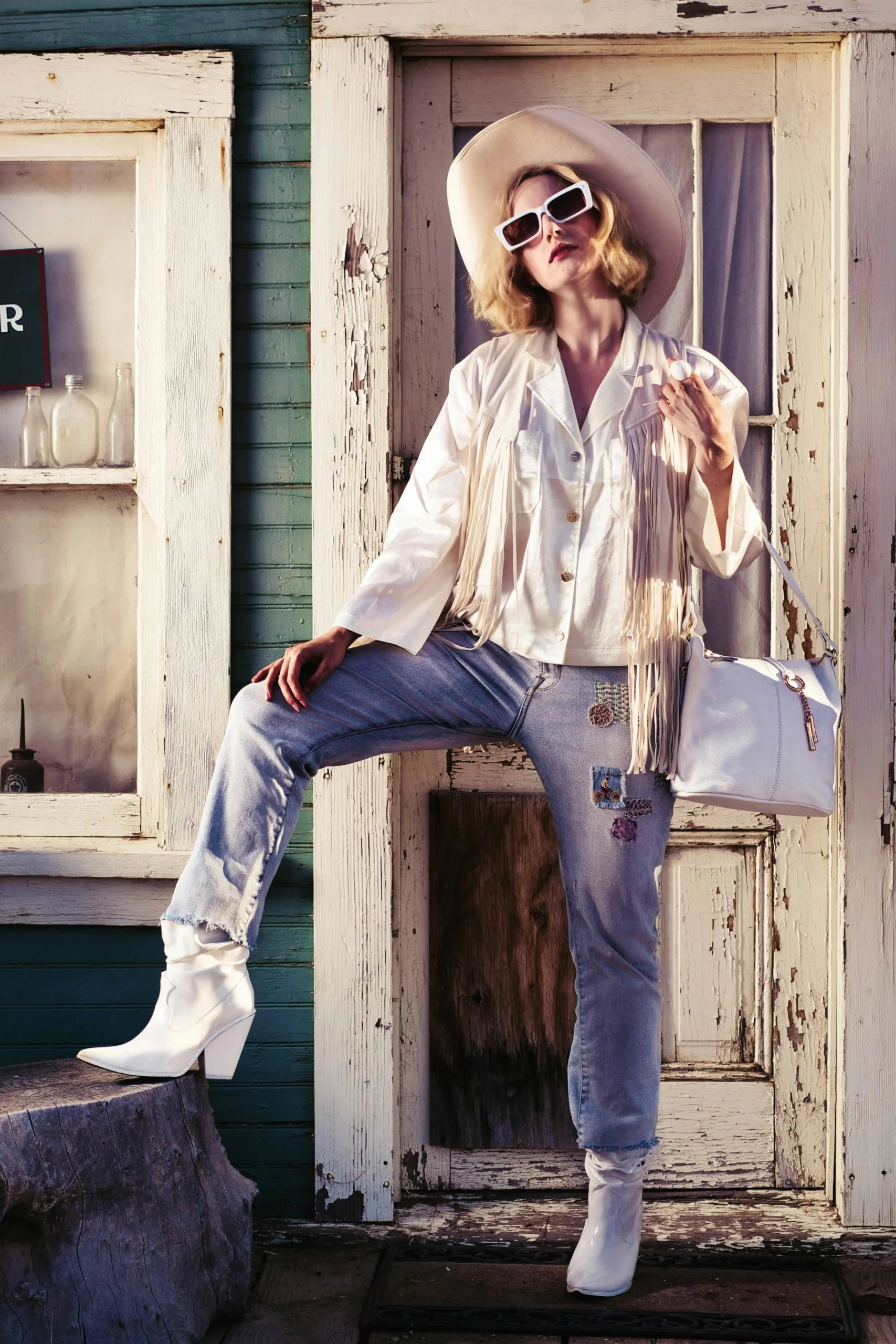 chic cowgirl confidently standing with one leg propped up in front of a chippy farmhouse door. Wearing white cowgirl boots and shirt and hat and purse, and light wash jeans.