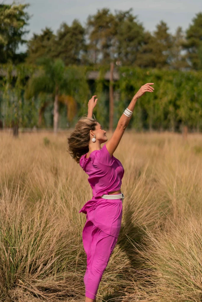 Stylish woman in fuschia outfit with bold white accessories, arms upstretched in the outdoors, appearing calm and joyful.