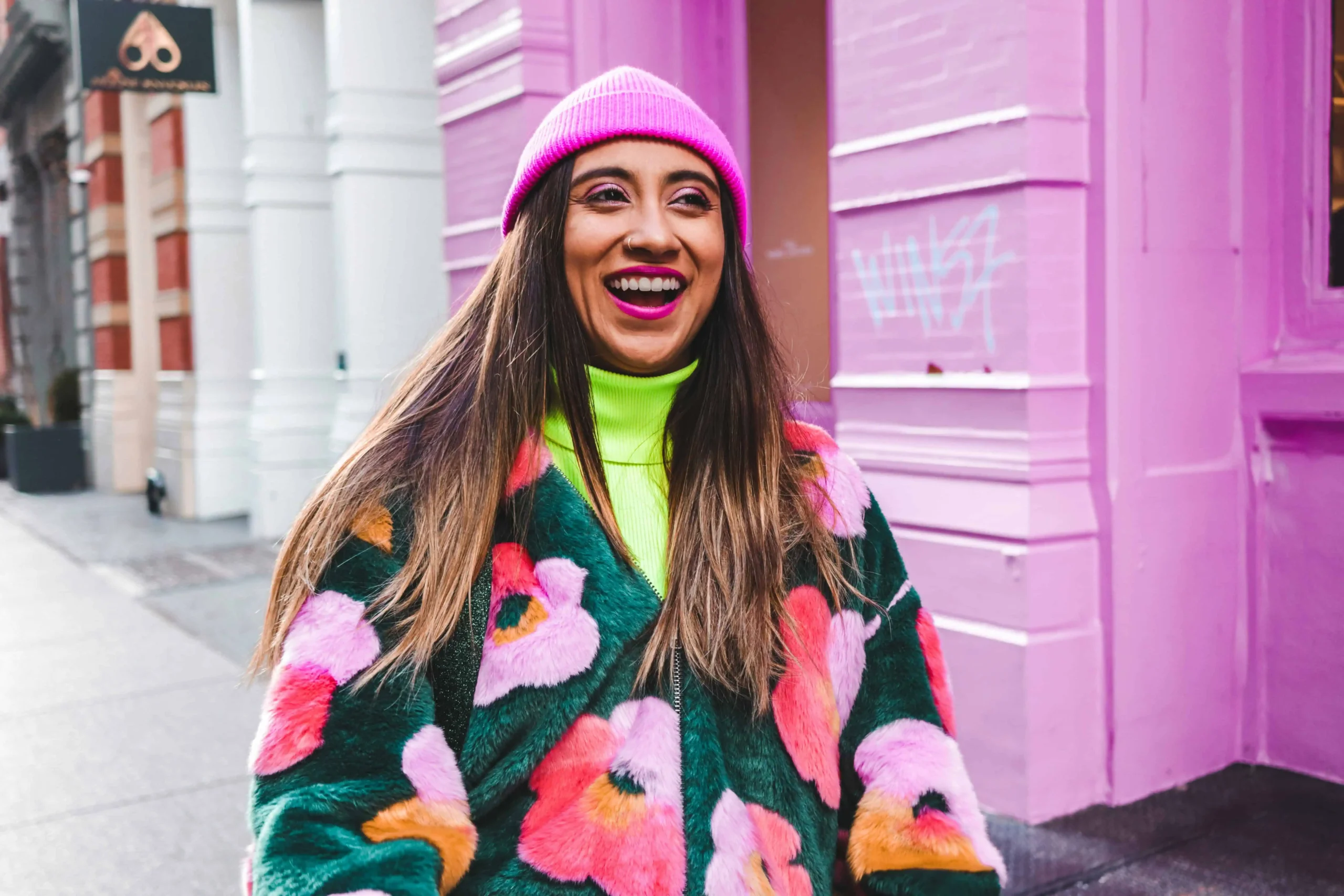 laughing young woman wearing floral fleece jacket with fuscia toque and a matching building in the background.