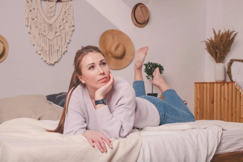 woman in neutral clothing laying on her bed with chin resting in her hand. Neutral decorations on wall in background.