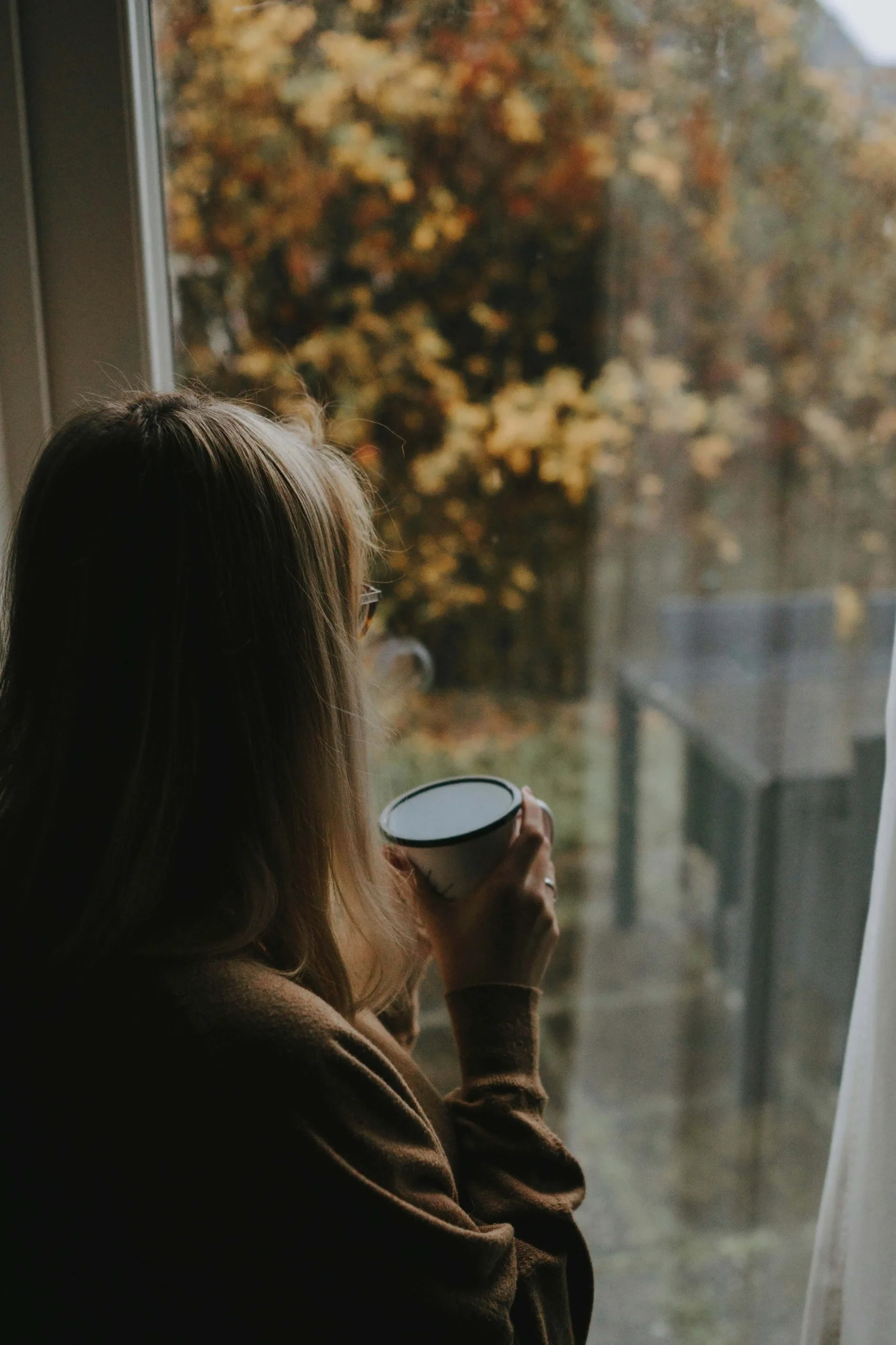 view from behind of woman holding coffee cup and staring out the window onto a drab leafy backyard