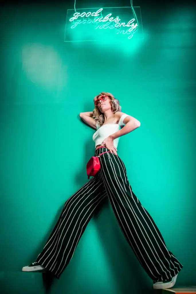 Trendy woman in black and white striped pants and converse shoes with brightly coloured pink belt purse, standing underneath a "good vibes only" sign. Deep vibrant teal green background.