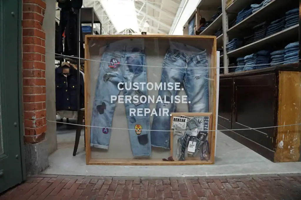 Sign for a store that does "Customise, Personalise & Repair" of clothing. It is actual jeans in a box, with lettering on the glass front encasing the display.